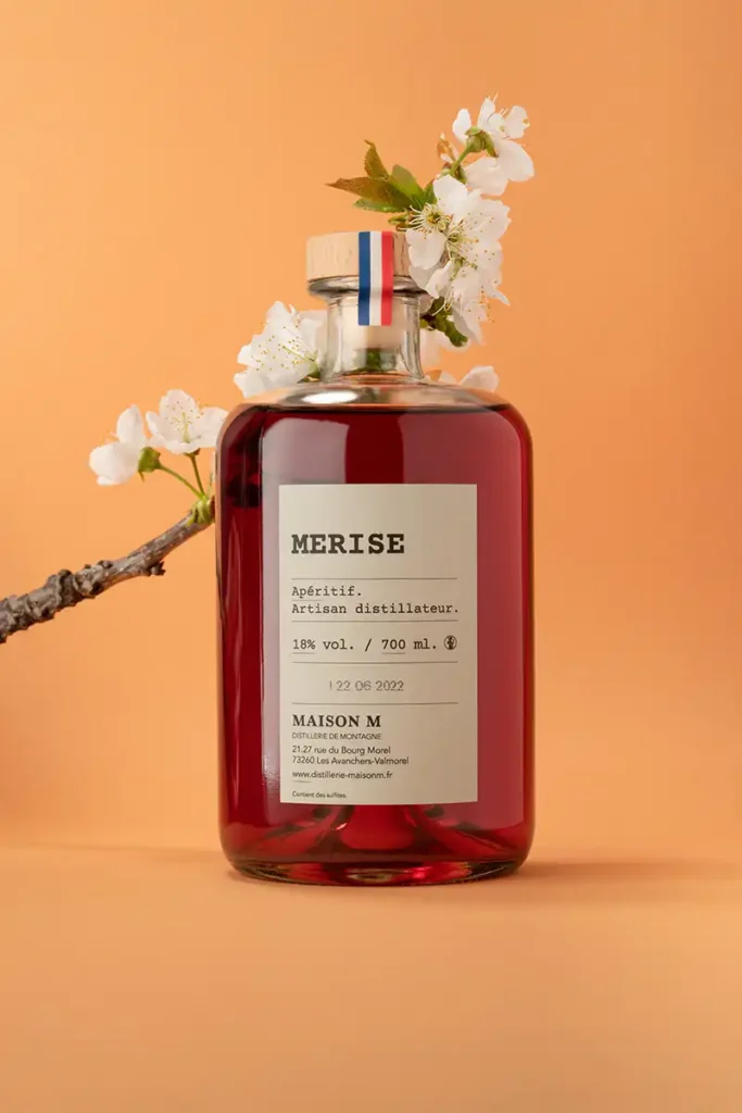 Flacon d'apéritif de montagne à la Merise de la distillerie Maison M avec des fleurs en arrière-plan.