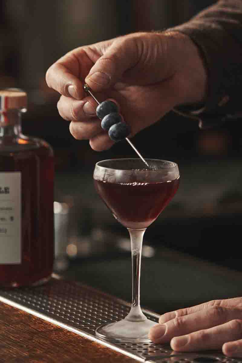 Photo d’un homme qui pose une brochette de myrtilles sur un cocktail servi dans un verre à pied